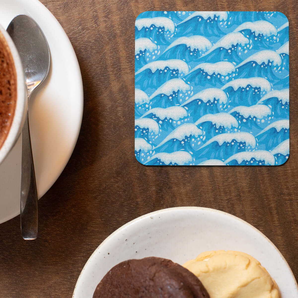 Decorative drink coaster with blue and white wave art next to a plate of cookies and a cup of coffee.