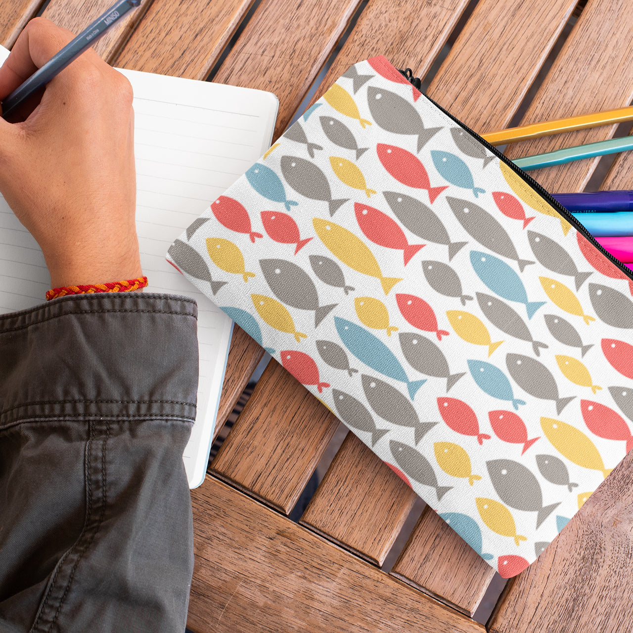 Woman writing in a notebook while using a colorful fish decor design pencil case pouch.