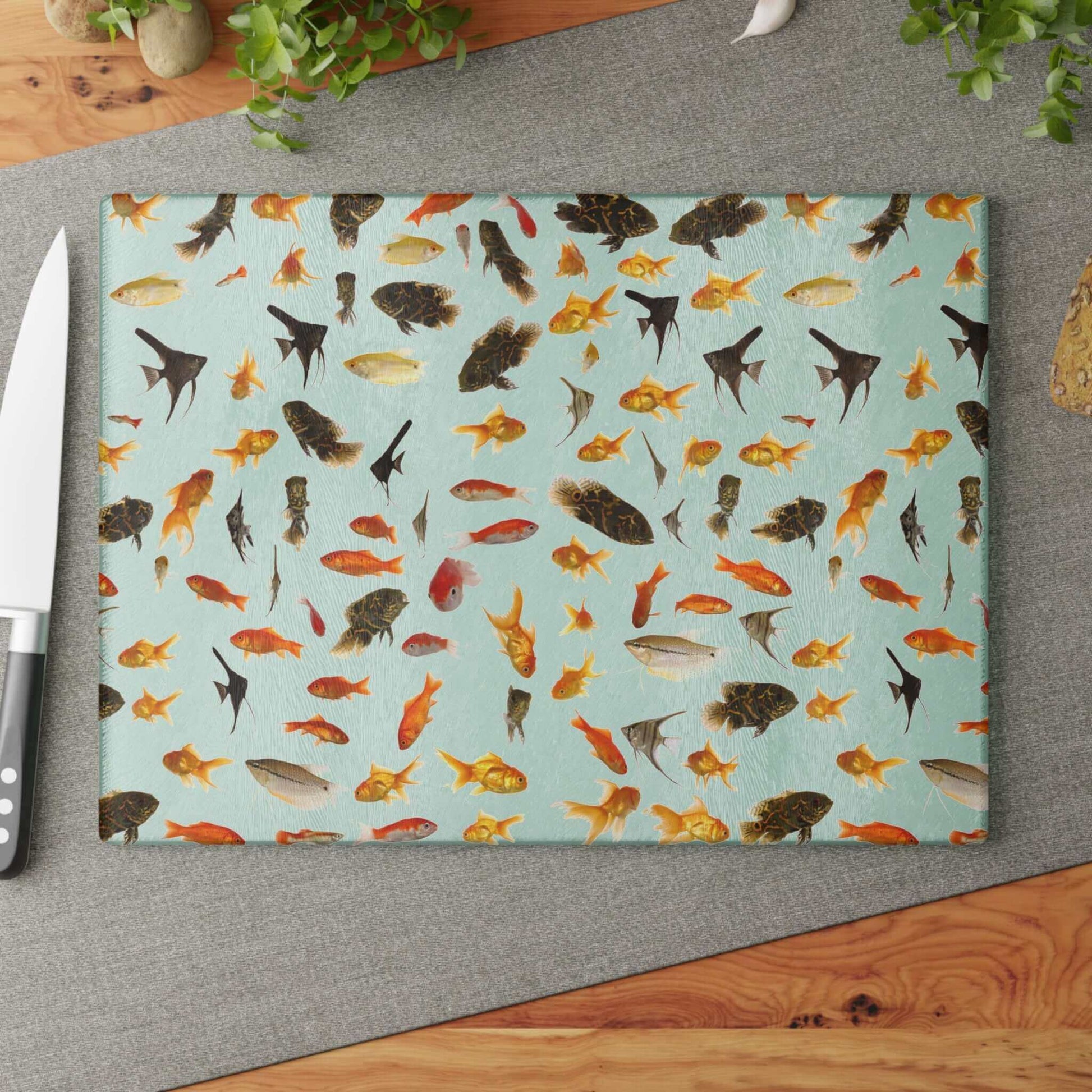 Colorful fish design on glass cutting board with knife and greenery nearby on a gray mat kitchen setup.