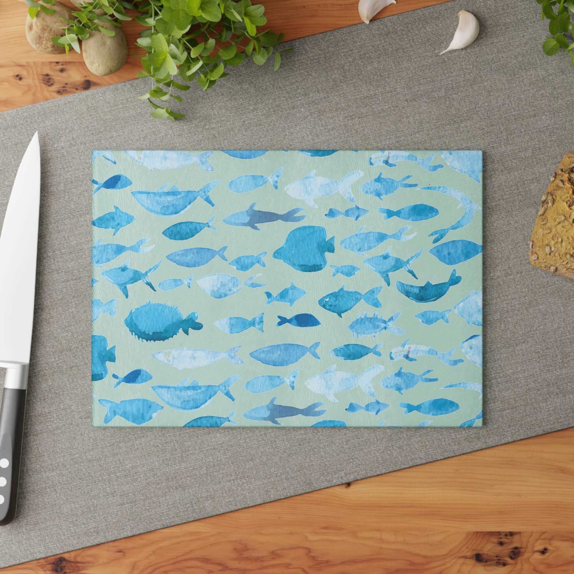 Blue watercolor fish design on durable glass cutting board, displayed on kitchen counter with knife and greenery