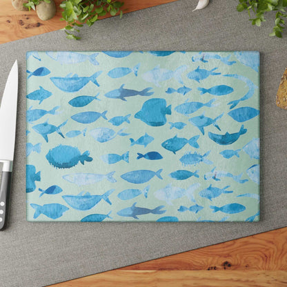 Blue watercolor fish glass cutting board on a gray mat with a knife, surrounded by fresh herbs.