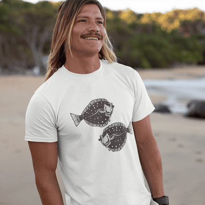 Man wearing Flounder T-Shirt with black fish design, standing on a beach.