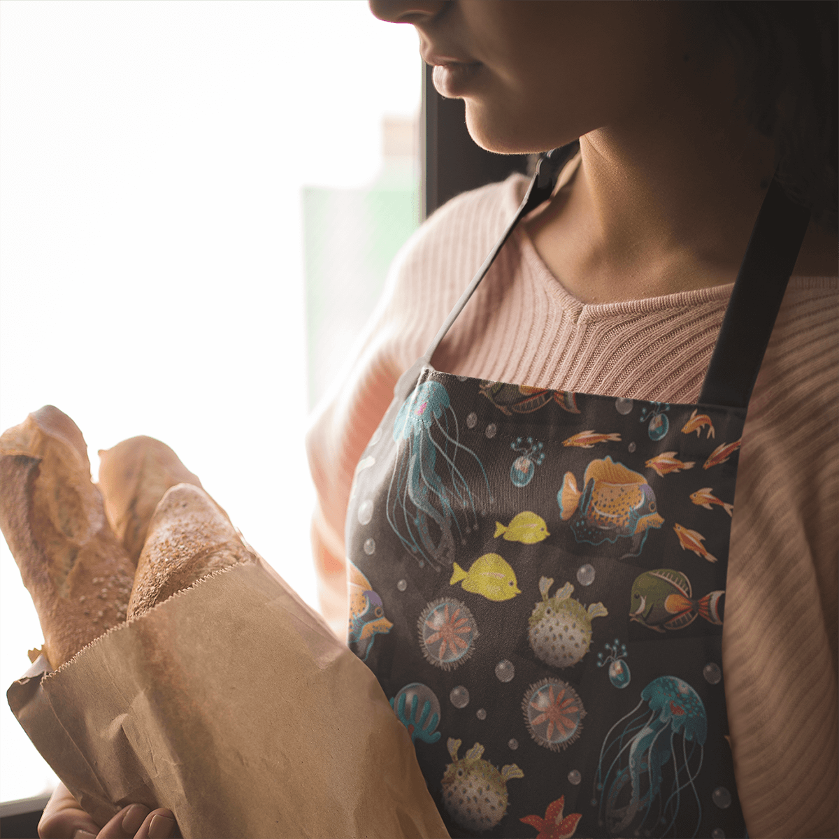 Woman wearing apron with deep sea fish and jellyfish design while holding a paper bag of fresh bread